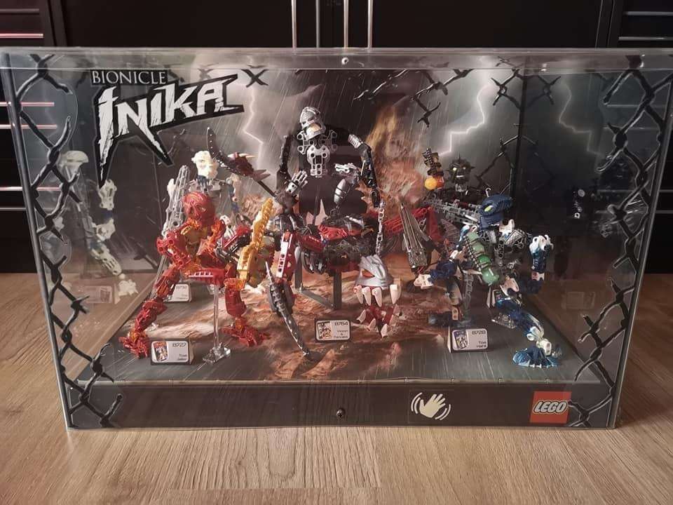 A large Lego display case featuring the Toa Inika Jaller, Hahli, Matoro, and Nuparu, along with the large Bionicle set "Vezon and Fenrakk". There is an icon towards the bottom right of the display that indicates a hand wave. The top left of the display has both the Bionicle and Inika logos. The corners of the display have a flat image of a broken chain-link fence